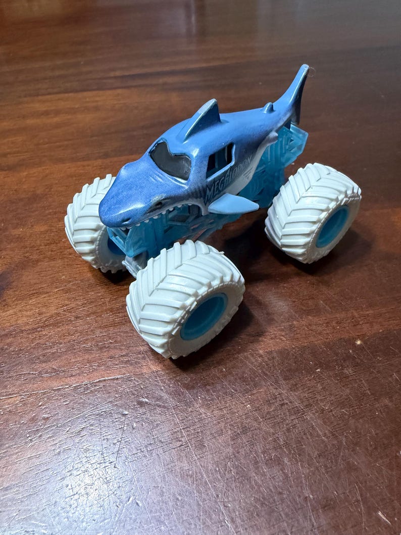 Puede incluir: Un cami&oacute;n monstruo de juguete azul y blanco con un dise&ntilde;o de tibur&oacute;n. El cami&oacute;n tiene neum&aacute;ticos blancos grandes con centros azules y la palabra "MEGAlodon" impresa en el lateral.