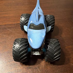 K&ouml;nnte beinhalten: Ein blaues Monstertruck-Spielzeug in Form eines Hais. Das Spielzeug hat gro&szlig;e schwarze Reifen und einen blauen K&ouml;rper mit einer Haifischflosse. Ein lustiges Spielzeug f&uuml;r Kinder.