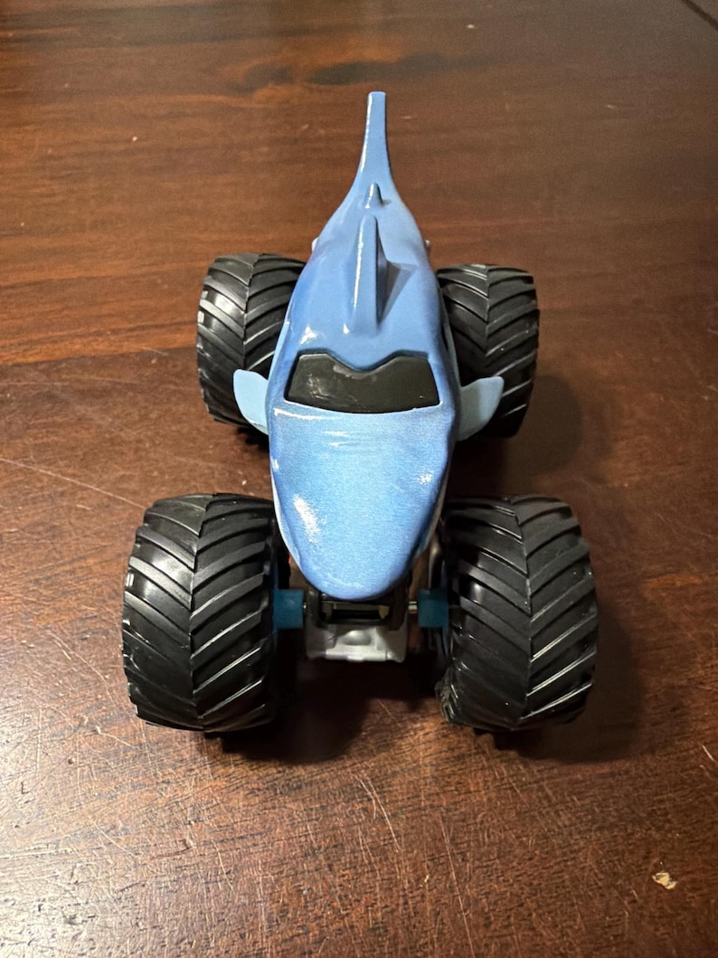 K&ouml;nnte beinhalten: Ein blaues Monstertruck-Spielzeug in Form eines Hais. Das Spielzeug hat gro&szlig;e schwarze Reifen und einen blauen K&ouml;rper mit einer Haifischflosse. Ein lustiges Spielzeug f&uuml;r Kinder.