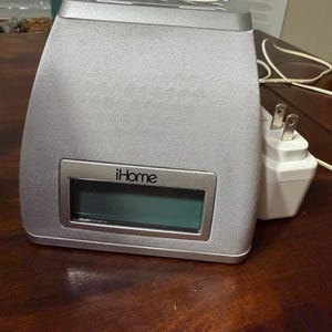 May include: Silver iHome alarm clock with a digital display. The clock has a rectangular display and a textured speaker surface. The power adapter is white with two prongs. The clock is on a dark brown wooden surface.