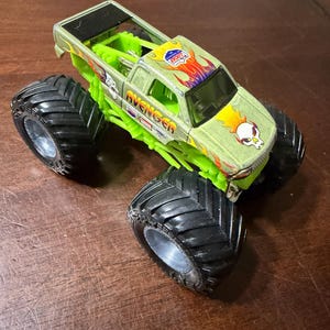 Puede incluir: Un cami&oacute;n monstruo de juguete verde y plateado con neum&aacute;ticos negros grandes. El cami&oacute;n presenta la palabra "AVENGER" y un gr&aacute;fico de calavera en el cap&oacute;. El cami&oacute;n tiene un chasis verde y un techo negro.