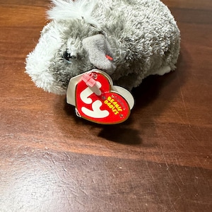 May include: A grey and white plush guinea pig toy with a red and white heart-shaped tag that reads "Beanie Babies". The toy has small black eyes and a tuft of white fur on its head. The tag is attached with a small red plastic piece.