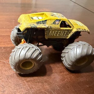 Peut inclure: Camion monstre jouet jaune et noir avec de gros pneus gris. Le camion porte l'inscription "MEENTS" sur le c&ocirc;t&eacute;. Le camion est pos&eacute; sur une surface marron.