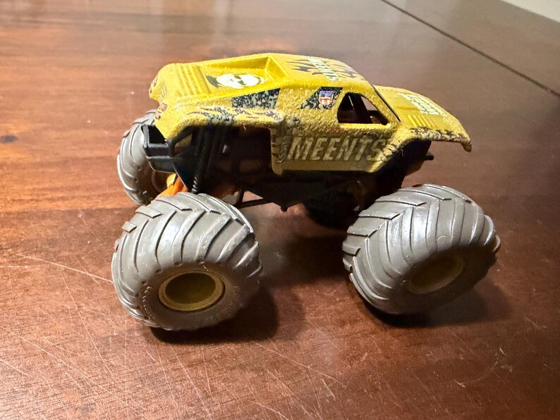 Peut inclure: Camion monstre jouet jaune et noir avec de gros pneus gris. Le camion porte l'inscription "MEENTS" sur le c&ocirc;t&eacute;. Le camion est pos&eacute; sur une surface marron.