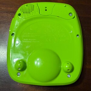 Pu&ograve; includere: Giocattolo elettronico verde brillante con un altoparlante, due pulsanti arrotondati e un grande pulsante circolare. Il retro del giocattolo &egrave; visibile, con la scritta "MADE IN CHINA" e diverse viti. Il giocattolo &egrave; su una superficie marrone scuro.