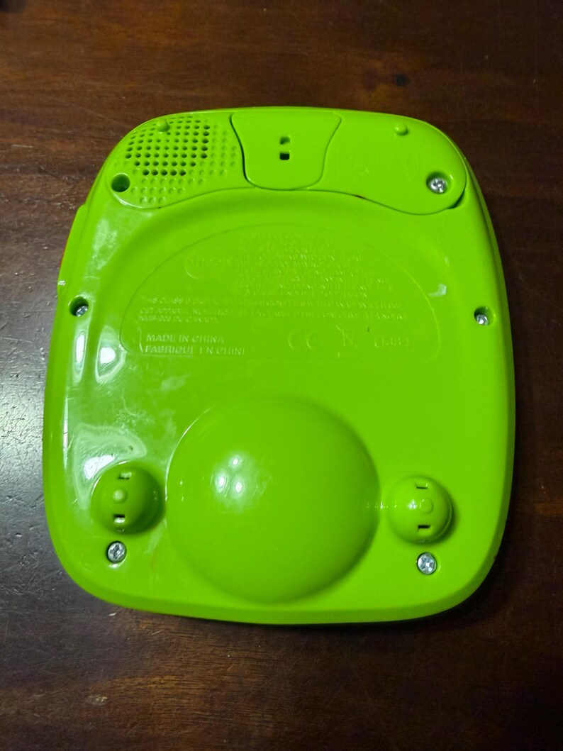 Pu&ograve; includere: Giocattolo elettronico verde brillante con un altoparlante, due pulsanti arrotondati e un grande pulsante circolare. Il retro del giocattolo &egrave; visibile, con la scritta "MADE IN CHINA" e diverse viti. Il giocattolo &egrave; su una superficie marrone scuro.