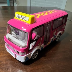Op de afbeelding: Een roze speelgoedbus met het woord "SNOOPY" op het dak en "PEANUTS" op een geel bord. De bus heeft heldere ramen, witte bekleding en cartoonafbeeldingen. De wielen zijn zilverkleurig. De bus staat op een bruin oppervlak.