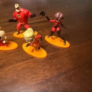 May include: A collection of The Incredibles character figurines. The figures are in dynamic poses, with Mr. Incredible, Elastigirl, and Violet visible. Each character stands on an orange oval base. The figures are primarily red and black.