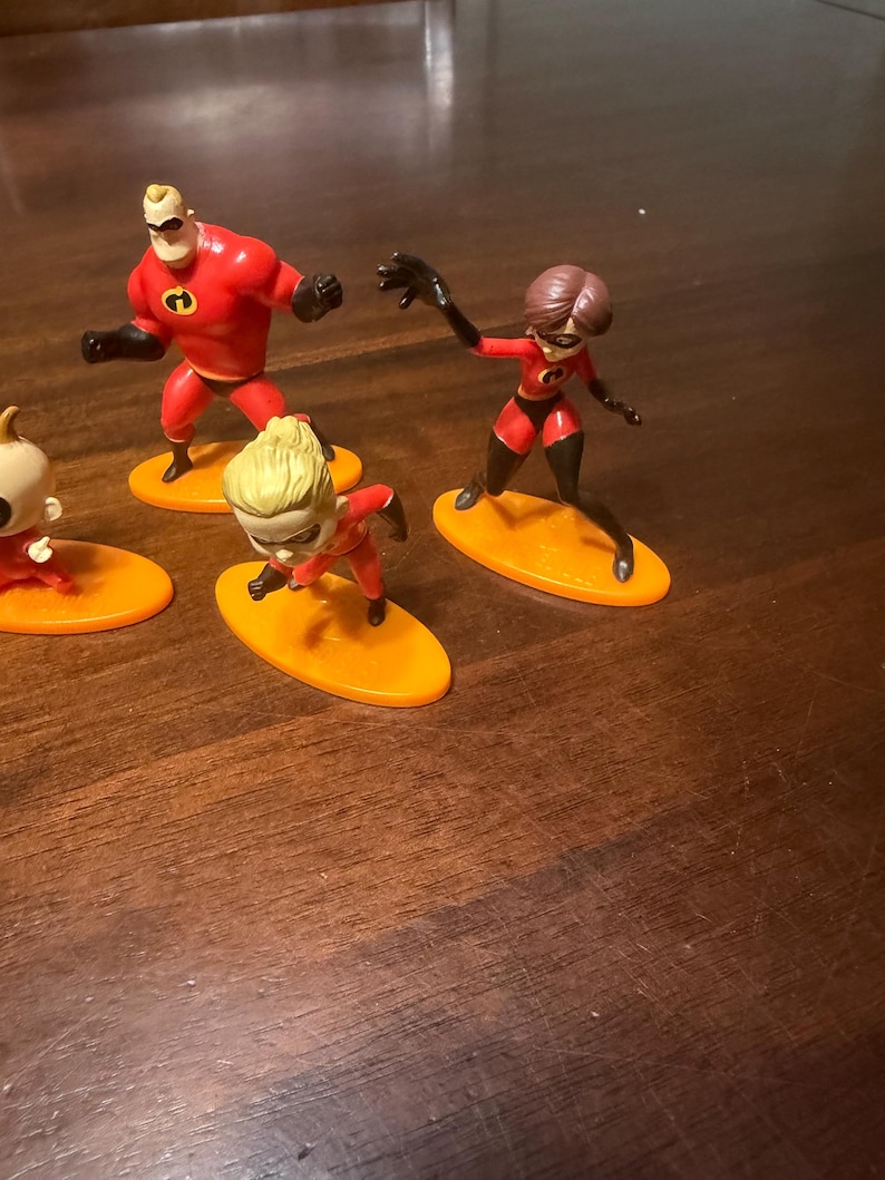May include: A collection of The Incredibles character figurines. The figures are in dynamic poses, with Mr. Incredible, Elastigirl, and Violet visible. Each character stands on an orange oval base. The figures are primarily red and black.