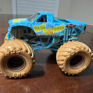May include: A light blue toy monster truck with large tan tyres. The truck has the words "BLUE THUNDER" in yellow and blue lightning bolt graphics. The truck is on a brown surface.