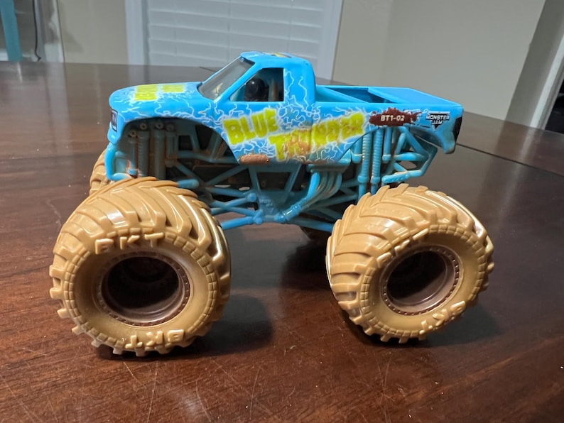 May include: A light blue toy monster truck with large tan tyres. The truck has the words "BLUE THUNDER" in yellow and blue lightning bolt graphics. The truck is on a brown surface.