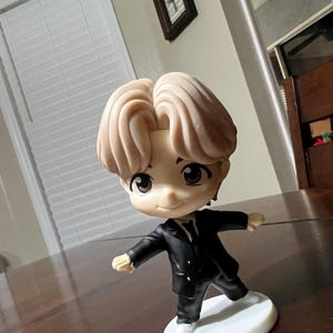 May include: A figurine of a person with light brown hair, wearing a black suit and white shoes, standing on a white base. The figurine has large, expressive eyes and a slight smile. The figure is posed with arms outstretched.