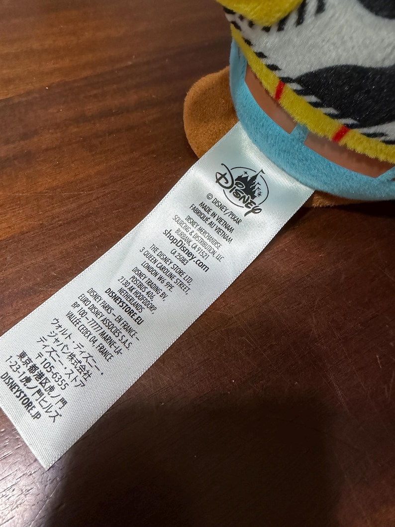 May include: A close-up of a white tag with the Disney logo and text. The tag reads "MADE IN VIETNAM" and includes the shopDisney.com website address. The tag also lists the Disney Store Ltd. address in London.