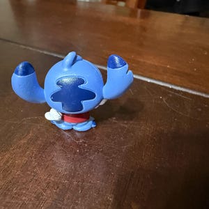 Pu&ograve; includere: Una statuetta blu di Stitch con grandi orecchie e un segno sulla testa blu scuro. La figura ha una camicia rossa e dettagli bianchi. La statuetta &egrave; su una superficie di legno marrone scuro.