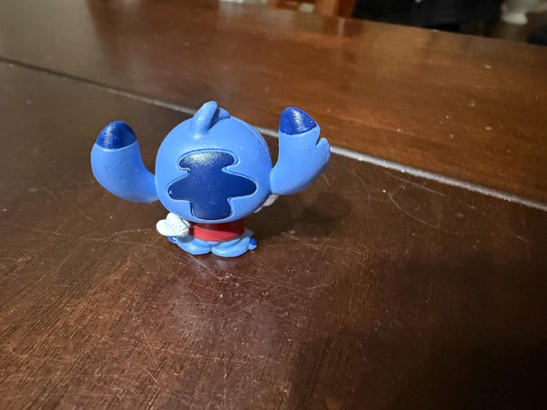 Pu&ograve; includere: Una statuetta blu di Stitch con grandi orecchie e un segno sulla testa blu scuro. La figura ha una camicia rossa e dettagli bianchi. La statuetta &egrave; su una superficie di legno marrone scuro.