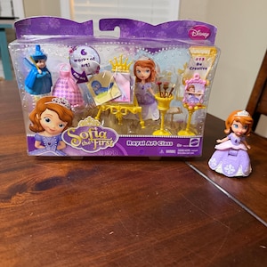 May include: A Disney Sofia the First Royal Art Class playset in a purple box. The set includes several small figurines, art supplies, and accessories. The box has the text "work of art!" and "#15 Be Creative". A separate Sofia figurine is on the table.