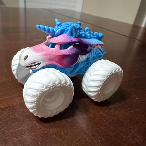 May include: A toy monster truck with a unicorn design. The truck has a white body, large white tyres, and a blue and pink unicorn head. The truck has a smiling face and a horn. The text "Unicorn SMASH" is visible on the side.