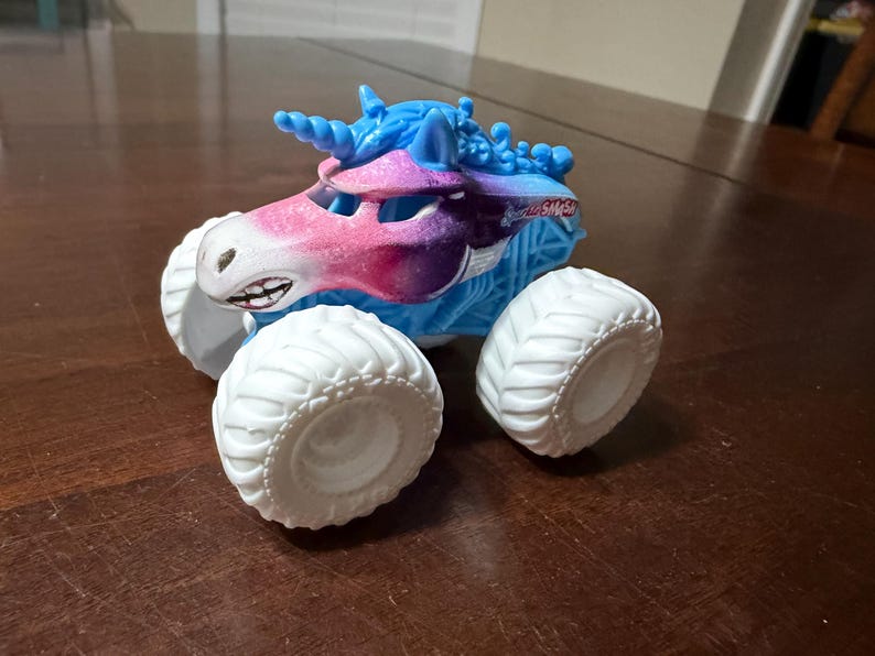 May include: A toy monster truck with a unicorn design. The truck has a white body, large white tyres, and a blue and pink unicorn head. The truck has a smiling face and a horn. The text "Unicorn SMASH" is visible on the side.
