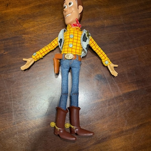 May include: A Woody action figure, featuring a yellow plaid shirt, blue jeans, and brown cowboy boots. The figure has a red bandana, a vest with a cow print, and a belt with a gold buckle. The toy's arms are outstretched.