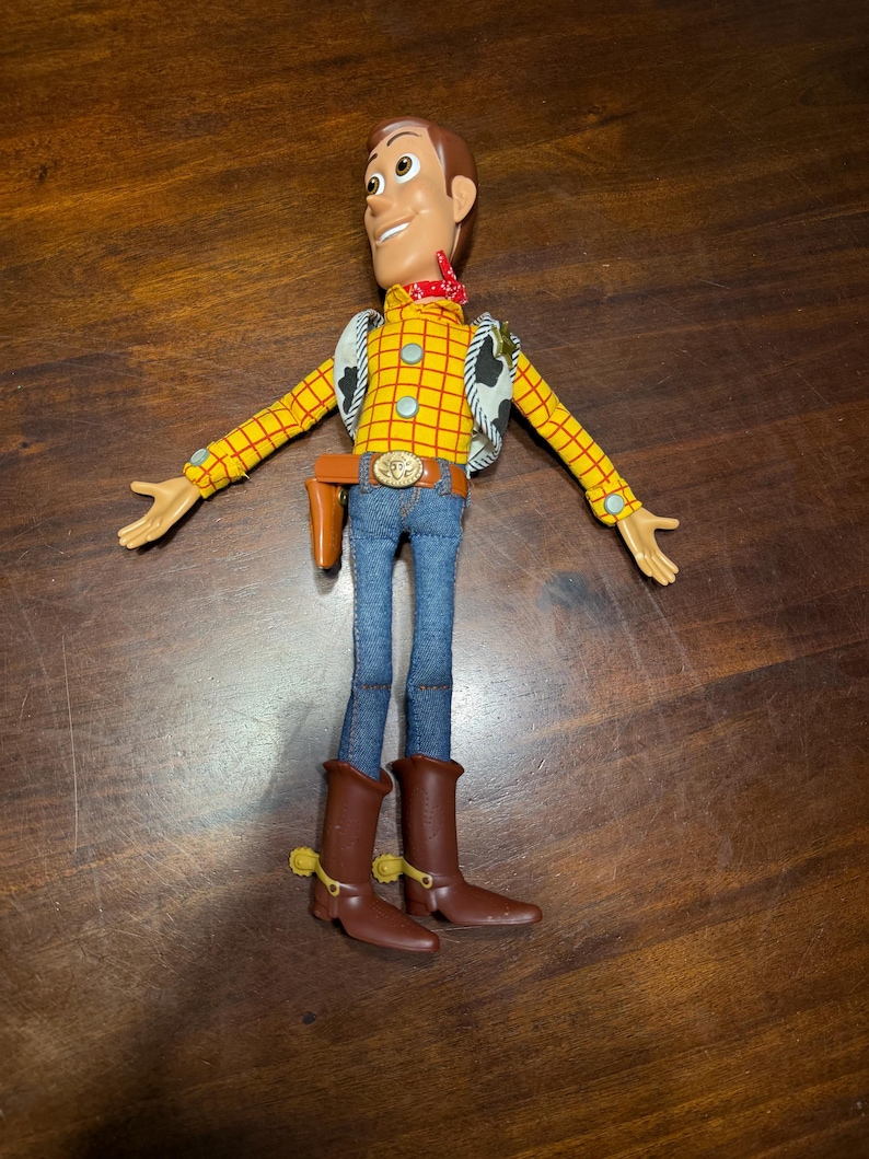 May include: A Woody action figure, featuring a yellow plaid shirt, blue jeans, and brown cowboy boots. The figure has a red bandana, a vest with a cow print, and a belt with a gold buckle. The toy's arms are outstretched.