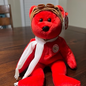 Puede incluir: Un oso de peluche rojo con gorro y gafas de aviador marrones, una bufanda blanca y un parche circular que dice "Too Worlds, Off Flight". El oso tiene ojos negros y nariz negra. Una etiqueta roja está adherida a la oreja del oso.