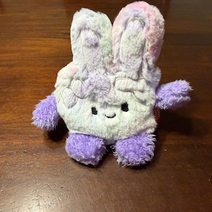 May include: A plush toy bunny in shades of purple, pink, and white. The bunny has long ears, a smiling face, and small arms and feet. The toy is made of soft, fuzzy material.