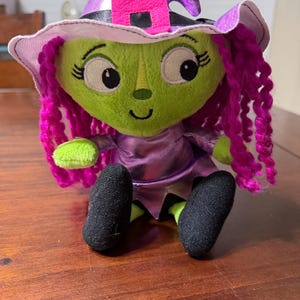 May include: A plush witch doll with a green face, pink curly hair, and a purple witch hat with a pink buckle. The doll wears a purple dress and black shoes. The doll is sitting on a wooden surface.