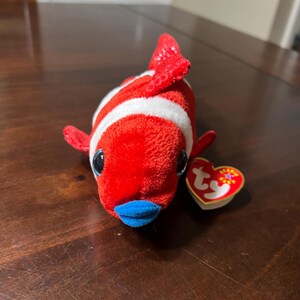 May include: Red and white striped plush fish with a blue mouth and black eyes. The fish has a white Ty tag attached to it.
