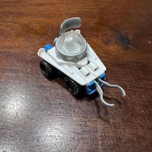 May include: A white toy vehicle with a clear dome, blue accents, and a silver claw arm. The vehicle has four wheels and a silver canopy.