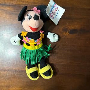 May include: A plush Minnie Mouse doll wearing a Hawaiian-themed outfit. The doll has a black body, white gloves, and yellow sandals. She wears a green grass skirt, a floral lei, and a pink flower on her head. A Disney Store tag is attached.