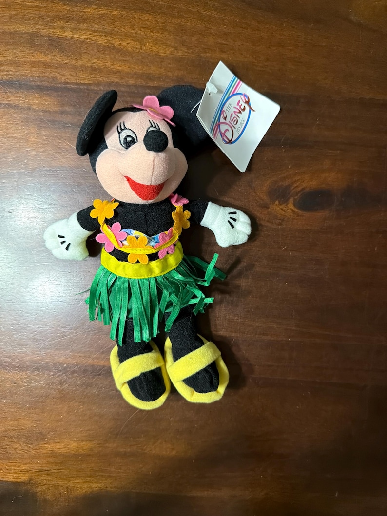 May include: A plush Minnie Mouse doll wearing a Hawaiian-themed outfit. The doll has a black body, white gloves, and yellow sandals. She wears a green grass skirt, a floral lei, and a pink flower on her head. A Disney Store tag is attached.