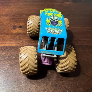 May include: A blue toy monster truck with large brown tires. The truck features a skull graphic with the words "Grave Digger" on the side. The top of the truck is green and yellow.