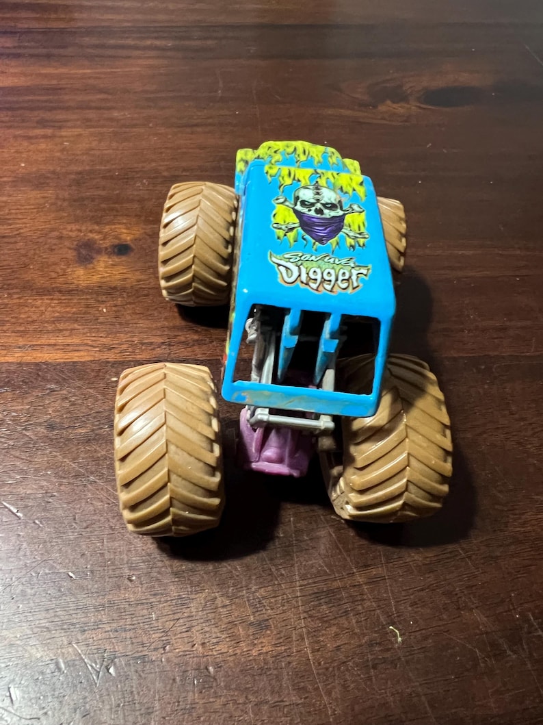 May include: A blue toy monster truck with large brown tires. The truck features a skull graphic with the words "Grave Digger" on the side. The top of the truck is green and yellow.