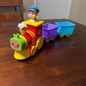 May include: A colorful toy train with a smiling watermelon face, red body, and yellow wheels. The train has a conductor figure and two attached cars, one purple and one blue. The toy is designed for children.