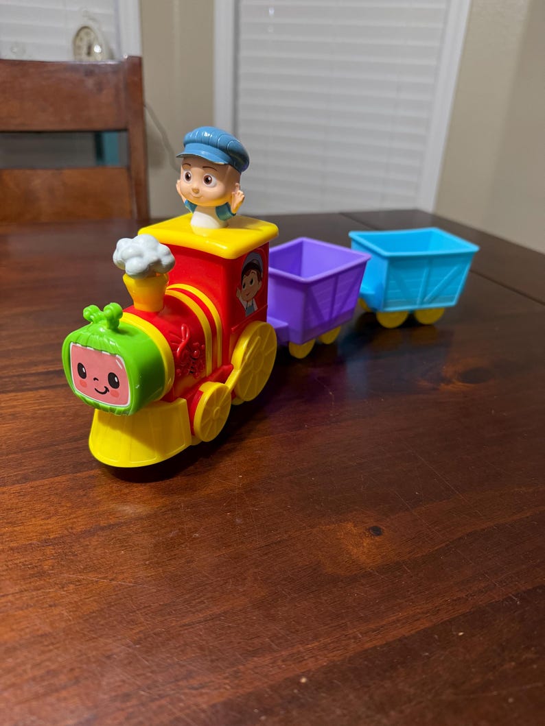 May include: A colorful toy train with a smiling watermelon face, red body, and yellow wheels. The train has a conductor figure and two attached cars, one purple and one blue. The toy is designed for children.