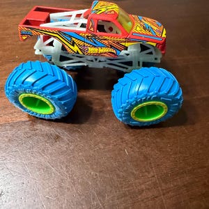 Puede incluir: Un cami&oacute;n monstruo Hot Wheels rojo, amarillo y azul con neum&aacute;ticos azules y llantas verdes. El cami&oacute;n tiene un dise&ntilde;o detallado con el logotipo de Hot Wheels y las palabras "Monster Trucks". Los neum&aacute;ticos est&aacute;n sueltos.