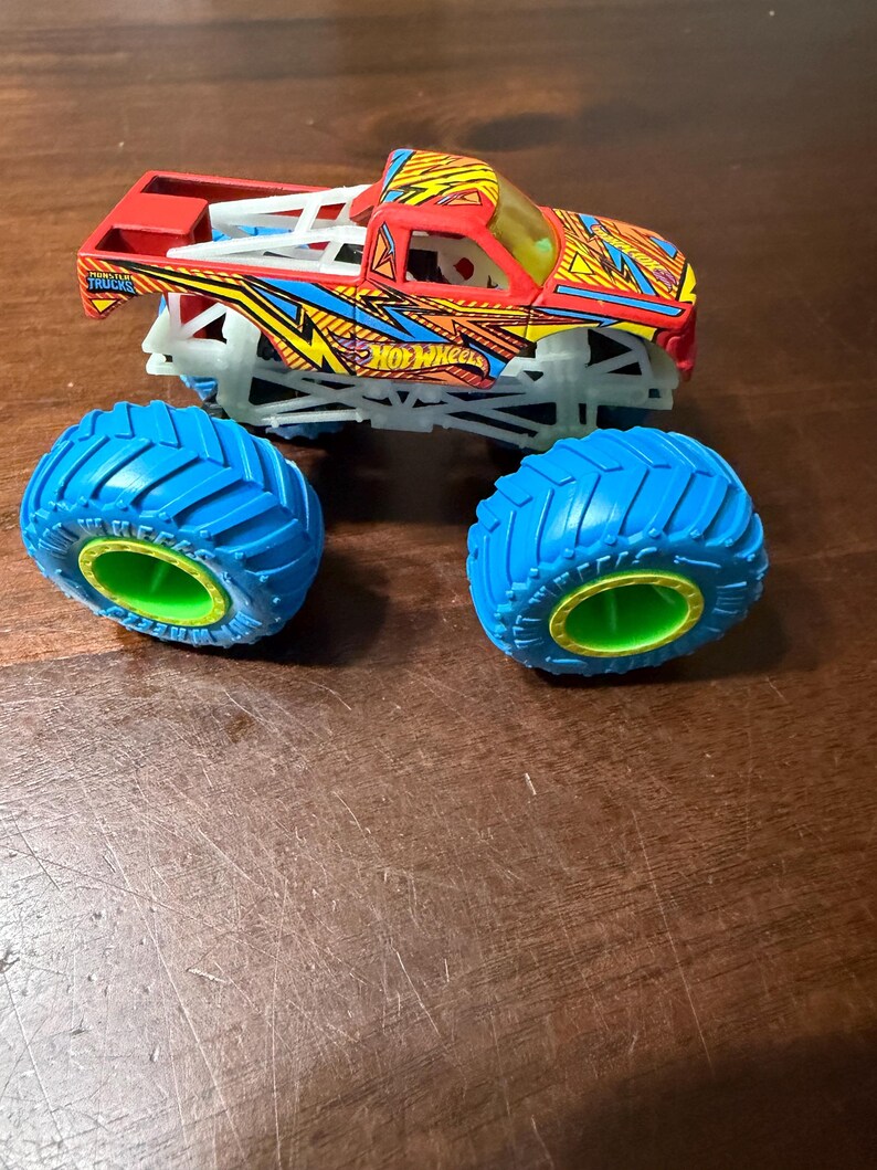 Puede incluir: Un cami&oacute;n monstruo Hot Wheels rojo, amarillo y azul con neum&aacute;ticos azules y llantas verdes. El cami&oacute;n tiene un dise&ntilde;o detallado con el logotipo de Hot Wheels y las palabras "Monster Trucks". Los neum&aacute;ticos est&aacute;n sueltos.