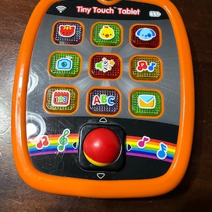 May include: A VTech Tiny Touch Tablet toy in orange and black. The tablet features nine colourful buttons with images, a red joystick, and rainbow accents. The text "Tiny Touch Tablet" is printed on the tablet.
