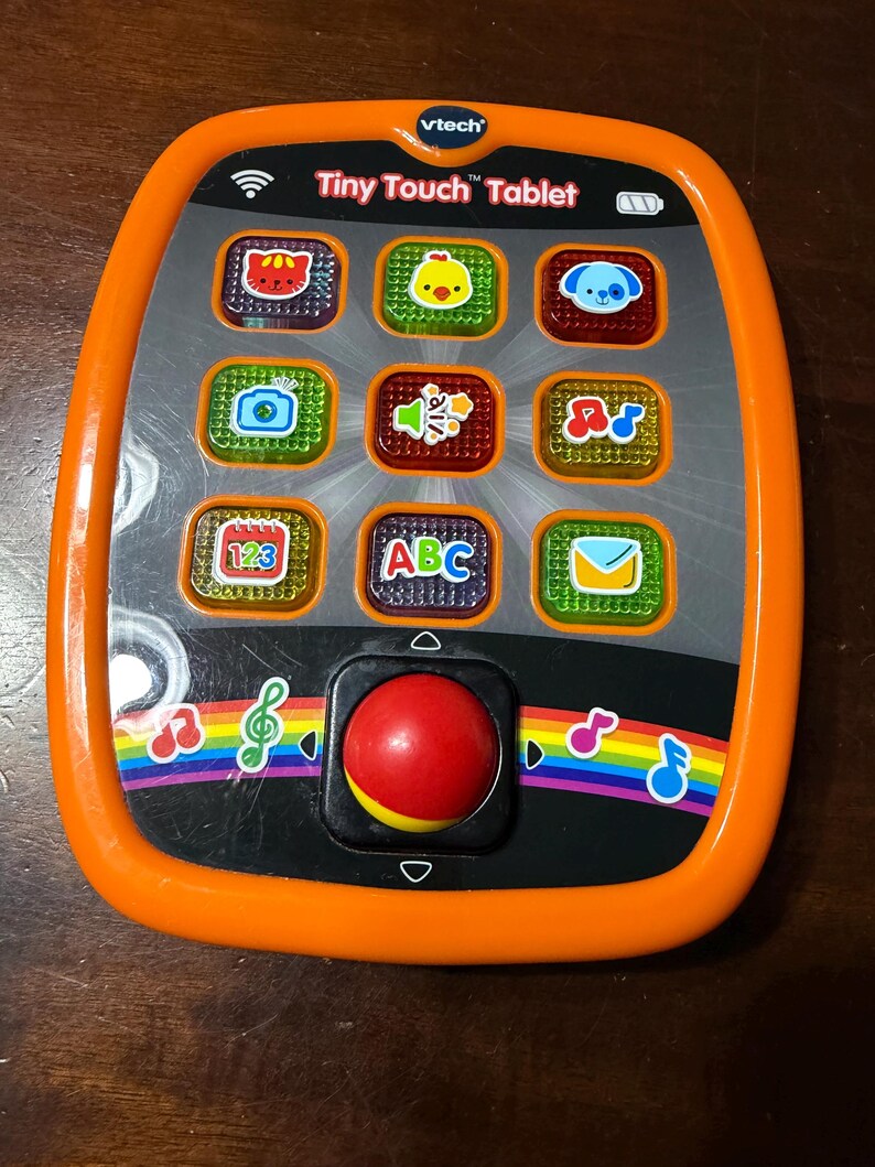Pu&ograve; includere: Un giocattolo VTech Tiny Touch Tablet arancione e nero. Il tablet ha nove pulsanti colorati con immagini, un joystick rosso e accenti arcobaleno. Il testo "Tiny Touch Tablet" &egrave; stampato sul tablet.