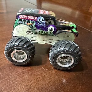 May include: A toy monster truck with large black tires and a chassis that glows in the dark. The truck is black with green and purple accents and the words "Grave Digger" on the roof.