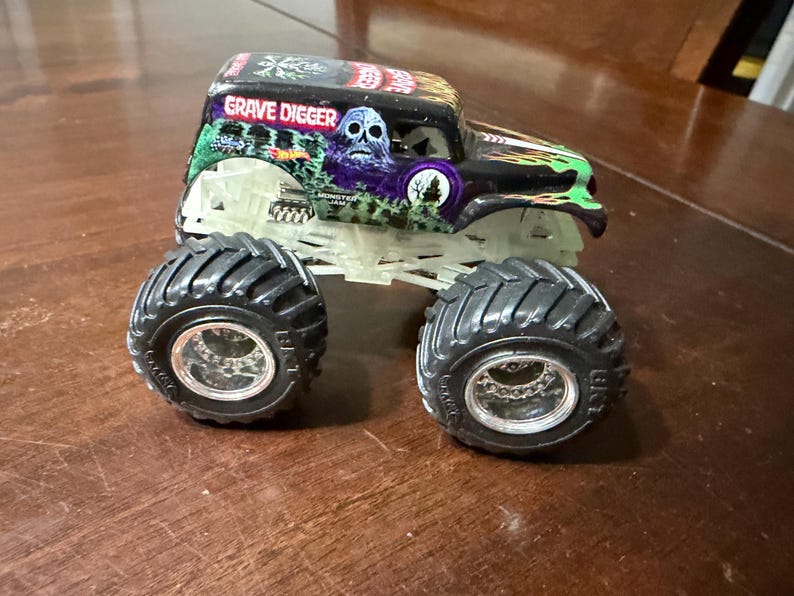 May include: A toy monster truck with large black tires and a chassis that glows in the dark. The truck is black with green and purple accents and the words "Grave Digger" on the roof.