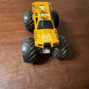 Puede incluir: Un cami&oacute;n monstruo de juguete amarillo con neum&aacute;ticos negros grandes. El cami&oacute;n tiene el texto "EMEENTS" y "2000-2001 WORLD CHAMPION" impreso. El cami&oacute;n tiene un dise&ntilde;o de llamas en los laterales.