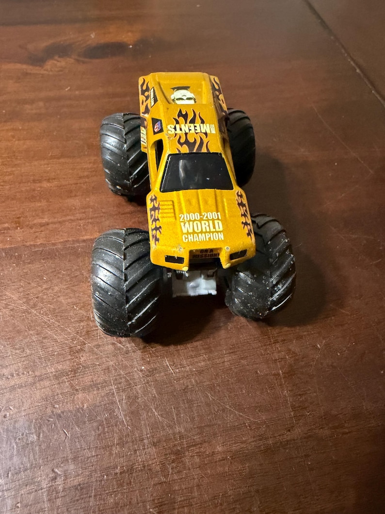 Puede incluir: Un cami&oacute;n monstruo de juguete amarillo con neum&aacute;ticos negros grandes. El cami&oacute;n tiene el texto "EMEENTS" y "2000-2001 WORLD CHAMPION" impreso. El cami&oacute;n tiene un dise&ntilde;o de llamas en los laterales.