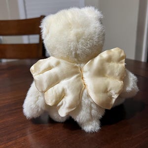 Puede incluir: Un oso de peluche color crema con alas de &aacute;ngel de sat&eacute;n. El oso est&aacute; sentado sobre una superficie de madera marr&oacute;n oscuro. Las alas son de color amarillo claro.