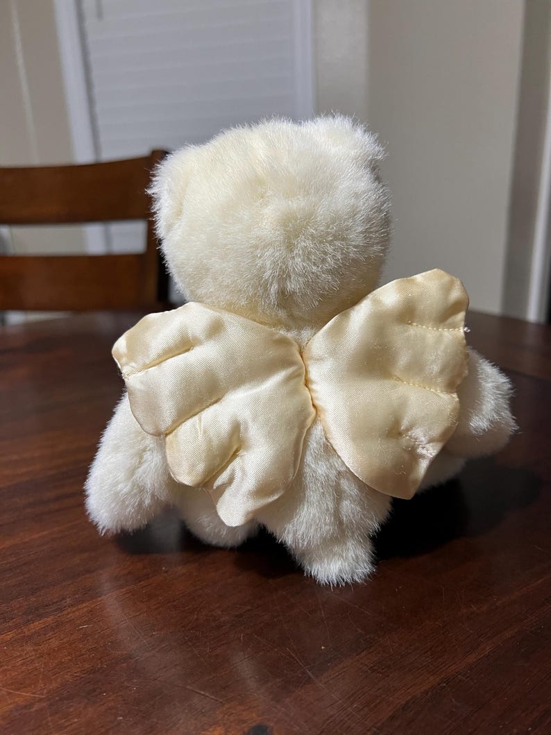 Puede incluir: Un oso de peluche color crema con alas de &aacute;ngel de sat&eacute;n. El oso est&aacute; sentado sobre una superficie de madera marr&oacute;n oscuro. Las alas son de color amarillo claro.