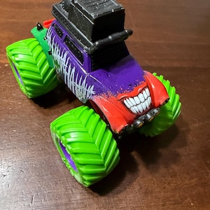 May include: A toy monster truck with a purple body, red front, and green wheels. The truck features a smiling face design and the word "HAHAHA" in white. A black structure sits on top.