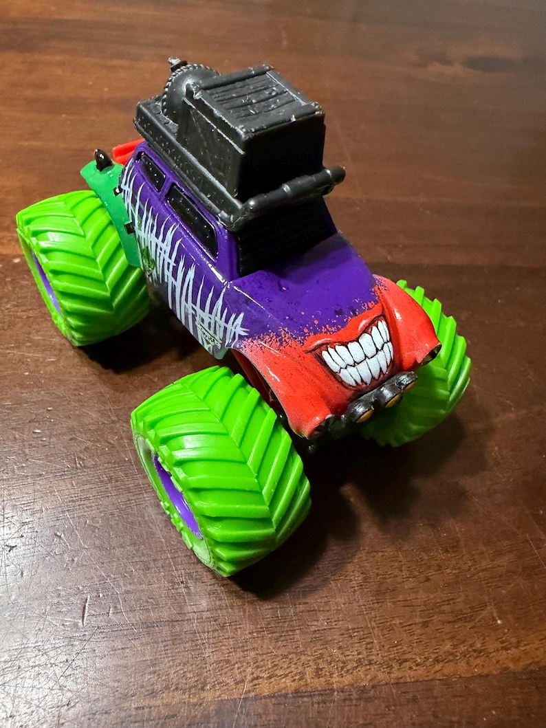 May include: A toy monster truck with a purple body, red front, and green wheels. The truck features a smiling face design and the word "HAHAHA" in white. A black structure sits on top.