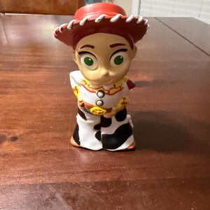 May include: A figurine of the character Jessie from the animated film Toy Story. The figurine has a red hat, a white shirt with red and yellow accents, a cow-print skirt, and a gold belt buckle. The character has green eyes and a smiling expression.