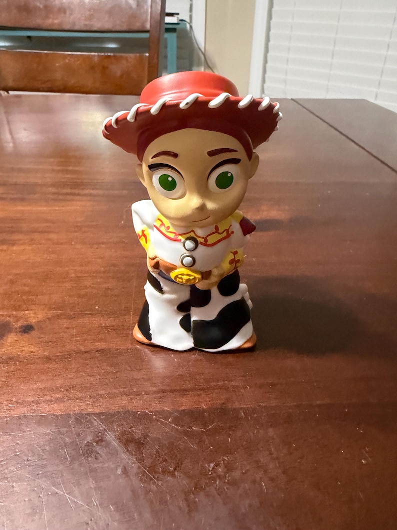 May include: A figurine of the character Jessie from the animated film Toy Story. The figurine has a red hat, a white shirt with red and yellow accents, a cow-print skirt, and a gold belt buckle. The character has green eyes and a smiling expression.