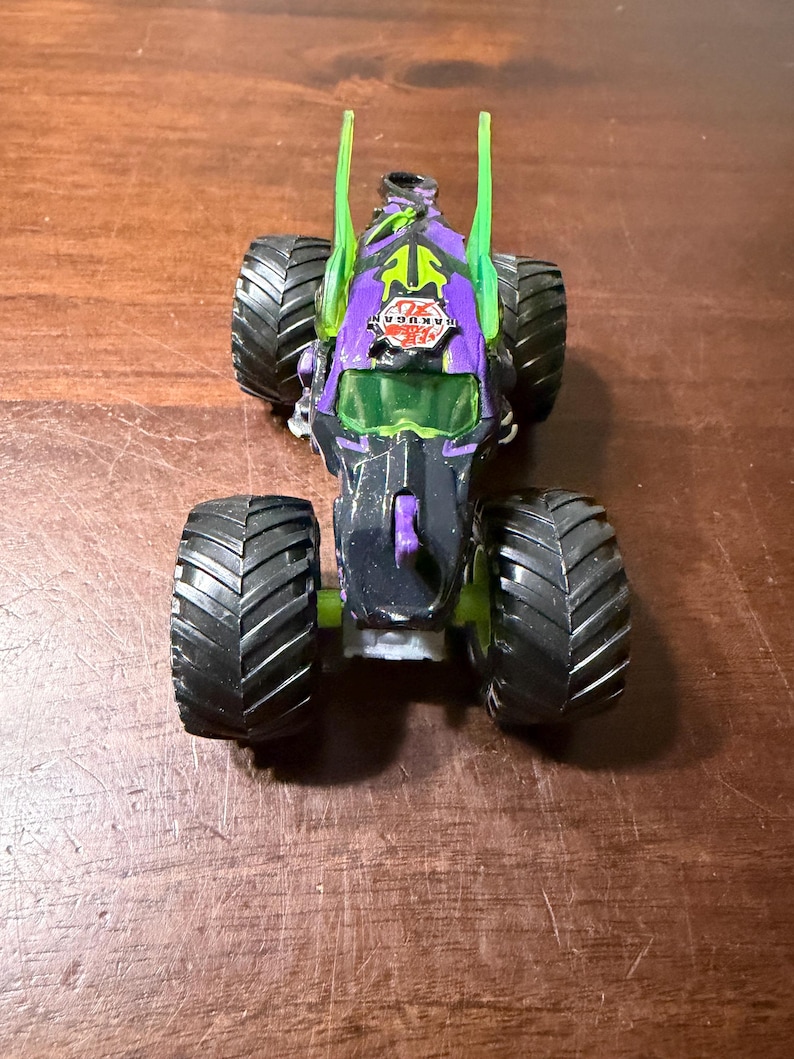 May include: A black and purple toy monster truck with large black tyres. The truck has green accents and the word "BAKUGAN" is visible on the front. The truck is sitting on a brown surface.